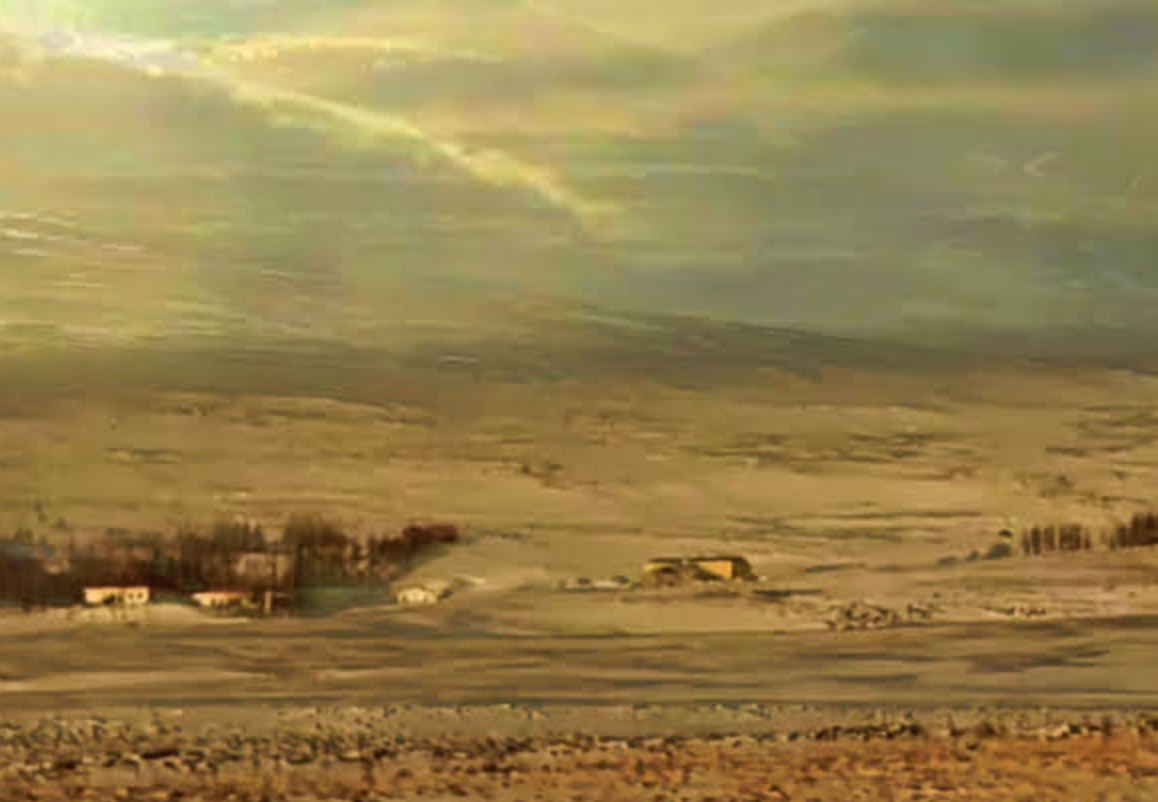 A glitched image of a beige landscape. The sky is full of clouds, but rays from the sun are visible between them. On the brown, blurry ground there are four small structures that could be houses. There is the suggestion of a mountain and trees in the background, but is it not clear. 