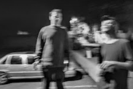 A blurry black and white image that appears to be a photograph. Two people, one wearing a hat, are stood near each other with a car in the background.