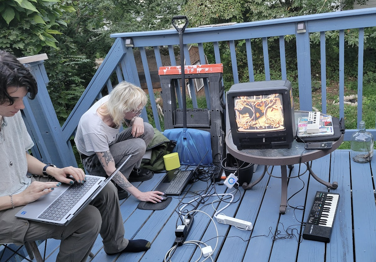 A photograph of two people working on laptops on a blue wooden porch, with foliage visible in the background. There is lots of equipment around them, such as an extension lead which is powering a small TV where it sits on a glass table, and a small keyboard which is on the floor. 