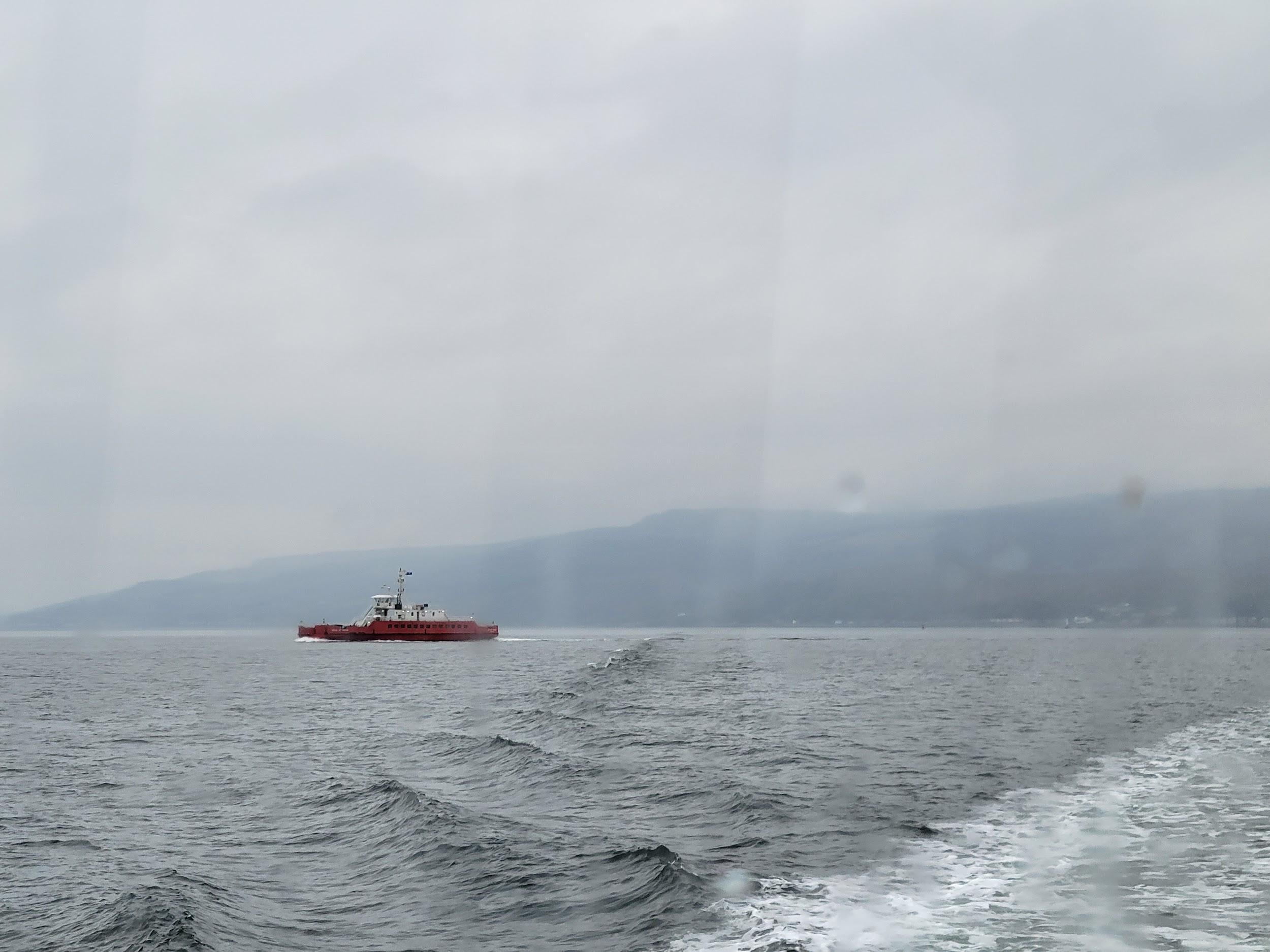 A photo of a red ferry at sea. It is a grey, foggy day, with a land mass visible behind the boat.