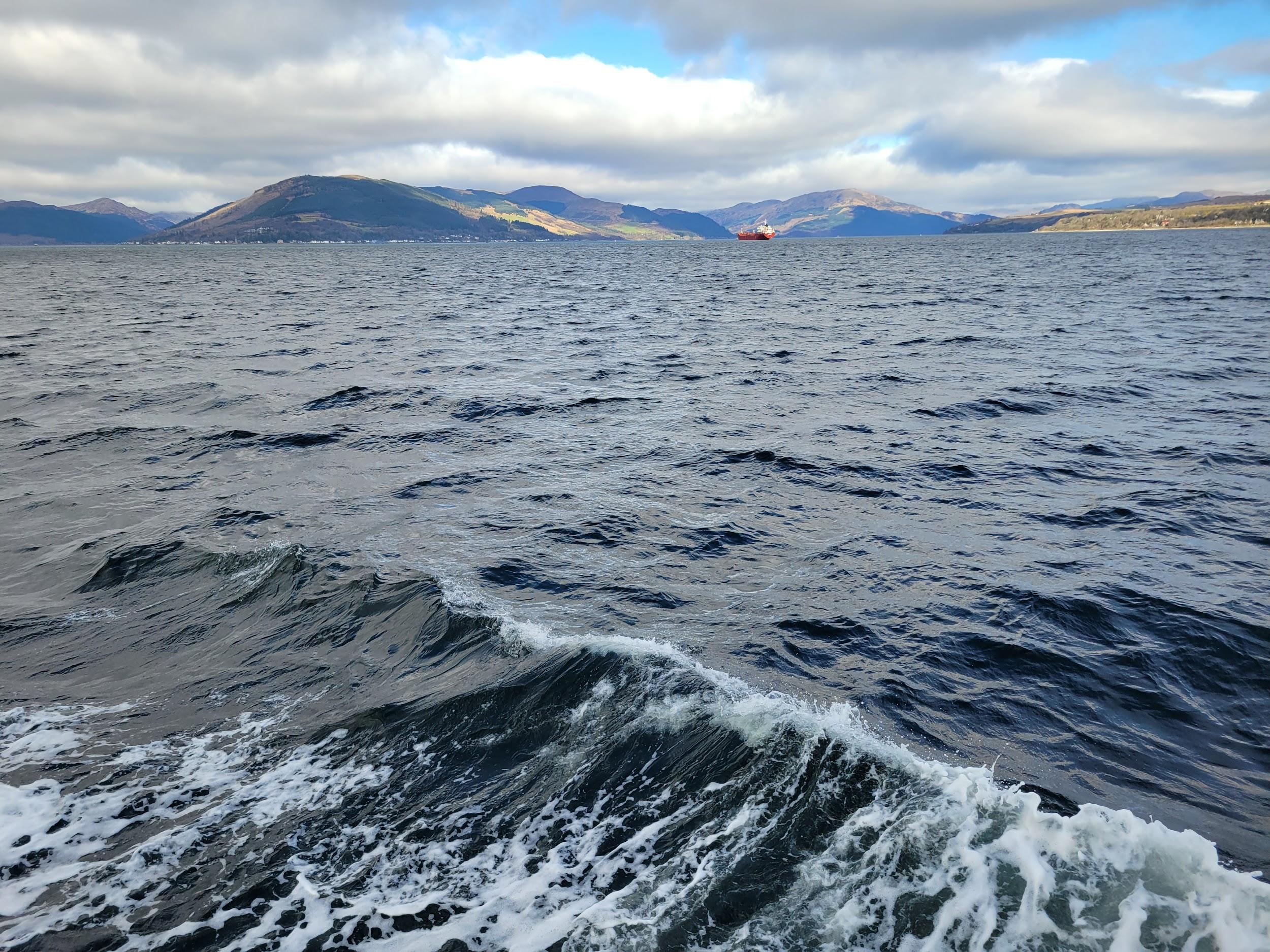 A photo of water, with hills and mountains visible on the distant shore. The sky is cloudy but there are patches of sunshine coming though. The only thing visible on the water is a red ferry.