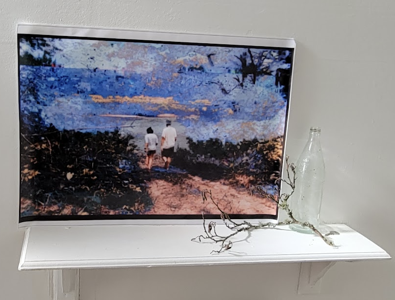 A printed landscape that is stuck to a wall above a shelf that has a small branch and empty glass bottle sat on it. The glitched landscape is of two people walking between bushes, as seen from behind. The sky is blue, with hints of gold.