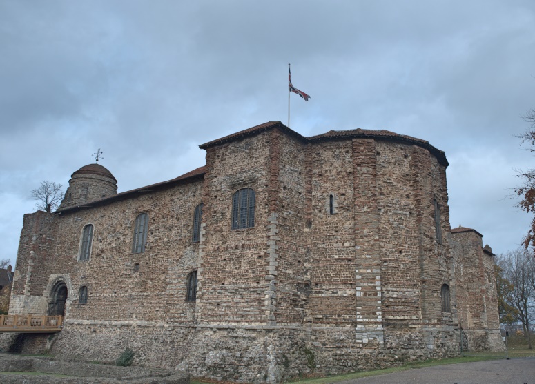 A stone building with a flag on top with Colchester Castle in the background